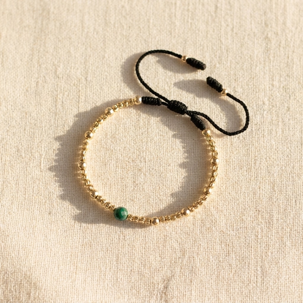 Pulsera fina con balines mini diamantados y esmeralda central oro laminado 18k sobre lino crema, flat lay editorial NJL Jewelry