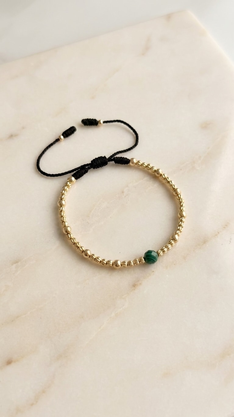 Pulsera fina con balines mini diamantados y esmeralda central oro laminado 18k flat lay sobre mármol cremoso, composición elegante NJL Jewelry